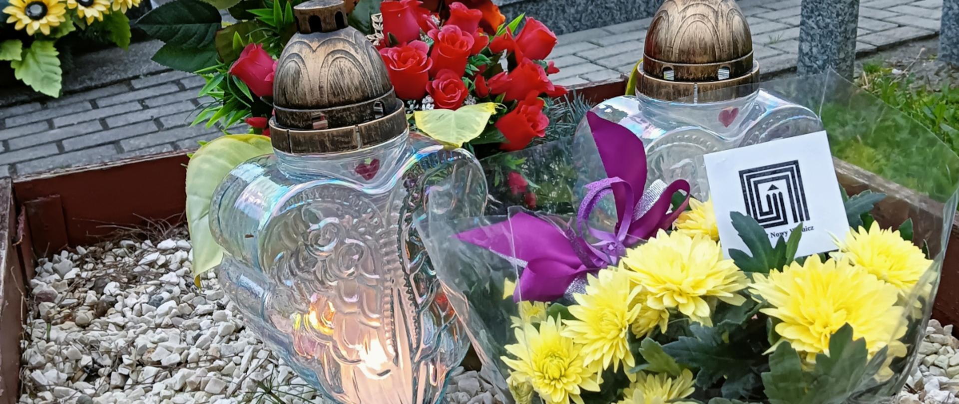fotografia barwna przedstawia kwiaty oraz ceramiczne i szklane znicze na drewnianym nagrobku wyłożonym białym kamieniem