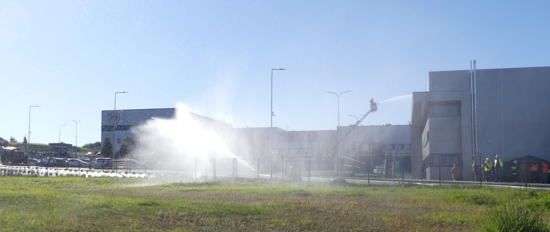 Zdjęcie przedstawia widok z oddali na budynki Wytwórni Opakowań Kartonowych w Brodnicy. Przed budynkami rozstawiony jest sprzęt pożarniczy z którego prowadzone a pozorowane działania gaśnicze. Woda leje się z poziomu ziemi – prawdopodobnie z prądownicy strażackiej oraz drugi prąd wody podawany jest z kosza podnośnika.