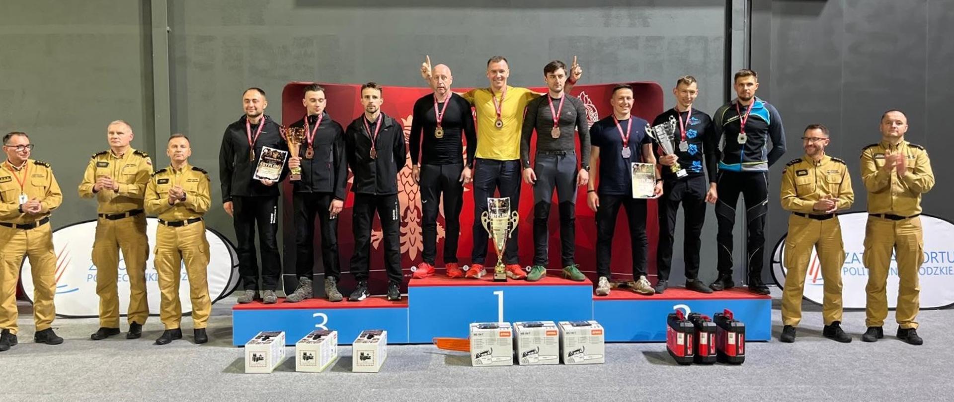 Dziewięciu zawodników w strojach sportowych na podium oraz pięciu strażaków w piaskowych mundurach galowych podczas dekoracji zwycięzców.