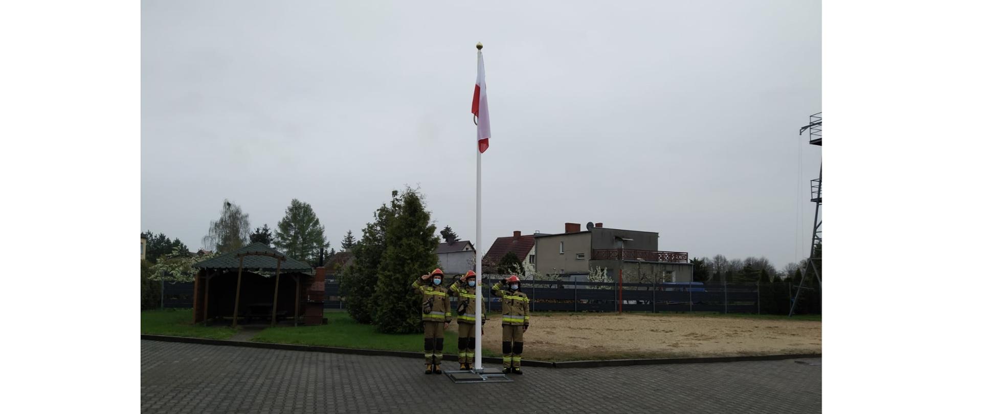 Fotografia przedstawia ujęcie placu z kostki polbrukowej. W centralnej części kadru stoi biały maszt, na nim zawieszona jest biało-czerwona flaga. Za masztem stoi trzech strażaków w ubraniach specjalnych, hełmach, z założonymi maseczkami chirurgicznymi, salutują. W tle widać tereny zielone jednostki ratowniczo-gaśniczej, dalej ogrodzenie i budynki mieszkalne. Jest dzień, niebo jest zachmurzone.