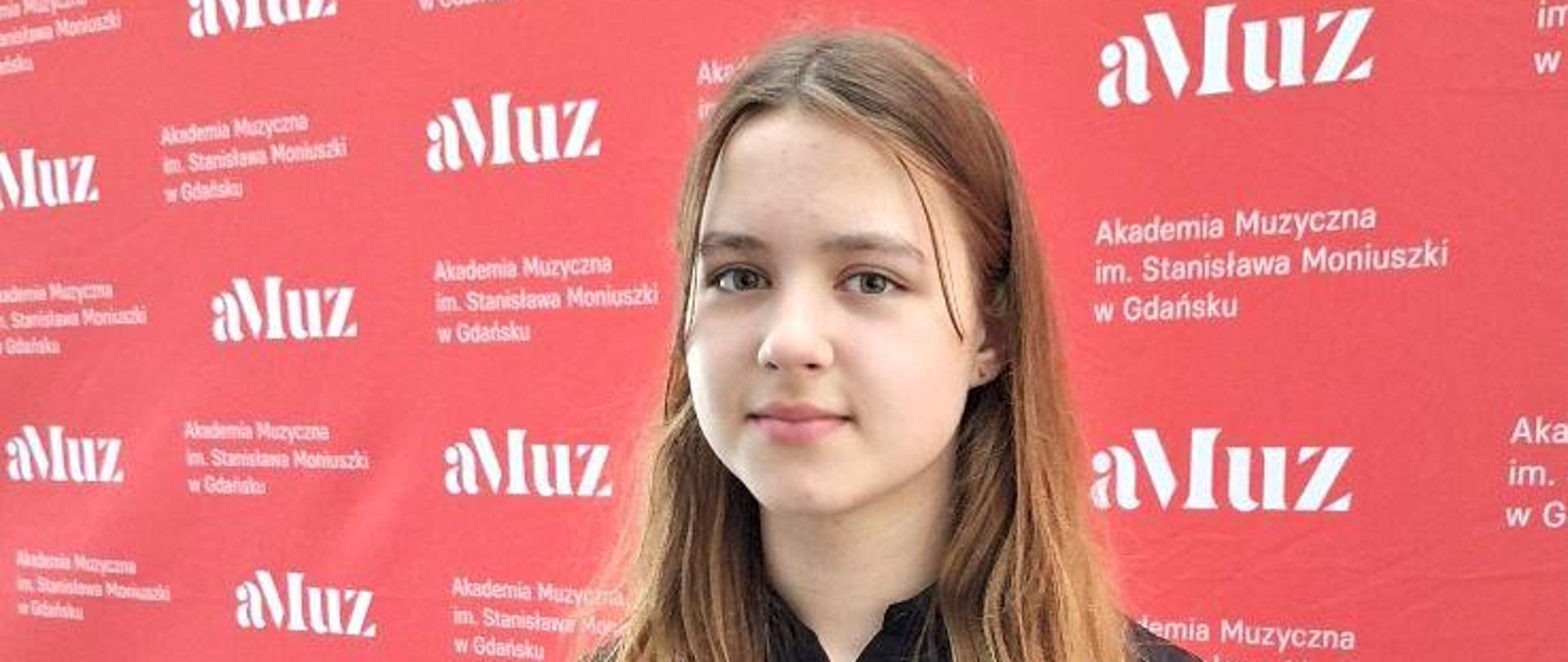 Na zdjęciu dziewczyna w długich włosach, ubrana w czarną sukienkę. Zdjęcie zostało wykonane na czerwonym tle z białymi napisami: "aMuz"