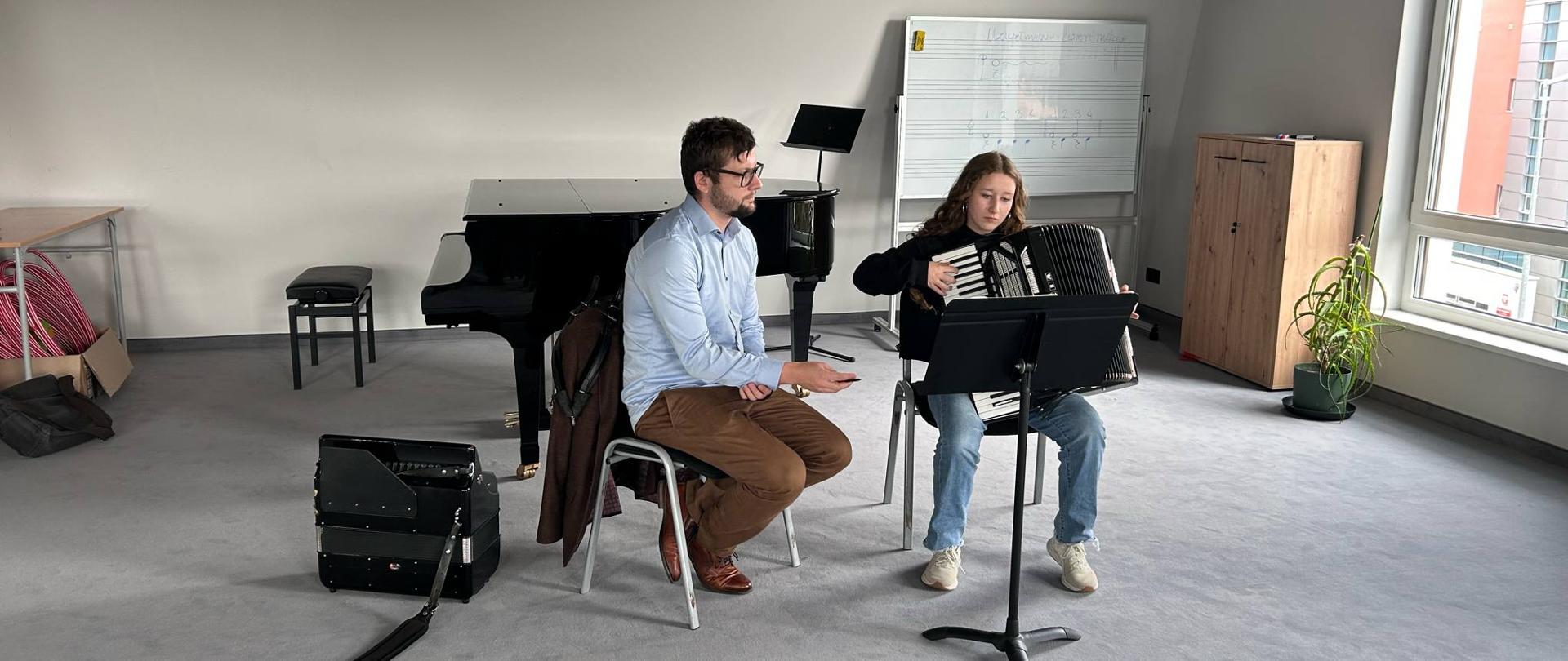 Na pierwszym planie znajduje się młoda osoba siedząca na krześle i grająca na akordeonie. Akordeon jest czarny i stoi na metalowym statywie z nutami. Osoba grająca ma długie, ciemne włosy spięte z tyłu głowy i ubrana jest w ciemny sweter z długim rękawem i jasne jeansy. Patrzy w dół na nuty. Za nią, po lewej stronie, siedzi prowadzący warsztaty na krześle. Jest zwrócony w stronę osoby grającej na akordeonie i wydaje się jej udzielać instrukcji. Mężczyzna ma krótkie, ciemne włosy i brodę. Ubrany jest w jasnoniebieską koszulę z długim rękawem i brązowe spodnie. Nosi okulary. W tle, po lewej stronie, stoi czarne pianino z otwartą klapą. Obok pianina stoi czarny stołek. Za pianinem znajduje się biała tablica z namalowanymi pięcioliniami. W prawym górnym rogu widać czarny głośnik zamontowany na ścianie. W prawym dolnym rogu stoi drewniana szafka, a obok niej duża roślina doniczkowa z zielonymi liśćmi. Za szafką znajduje się okno, przez które widać budynki. Pomieszczenie ma jasnoszare ściany i podłogę. Jest dobrze oświetlone światłem naturalnym wpadającym przez okno.