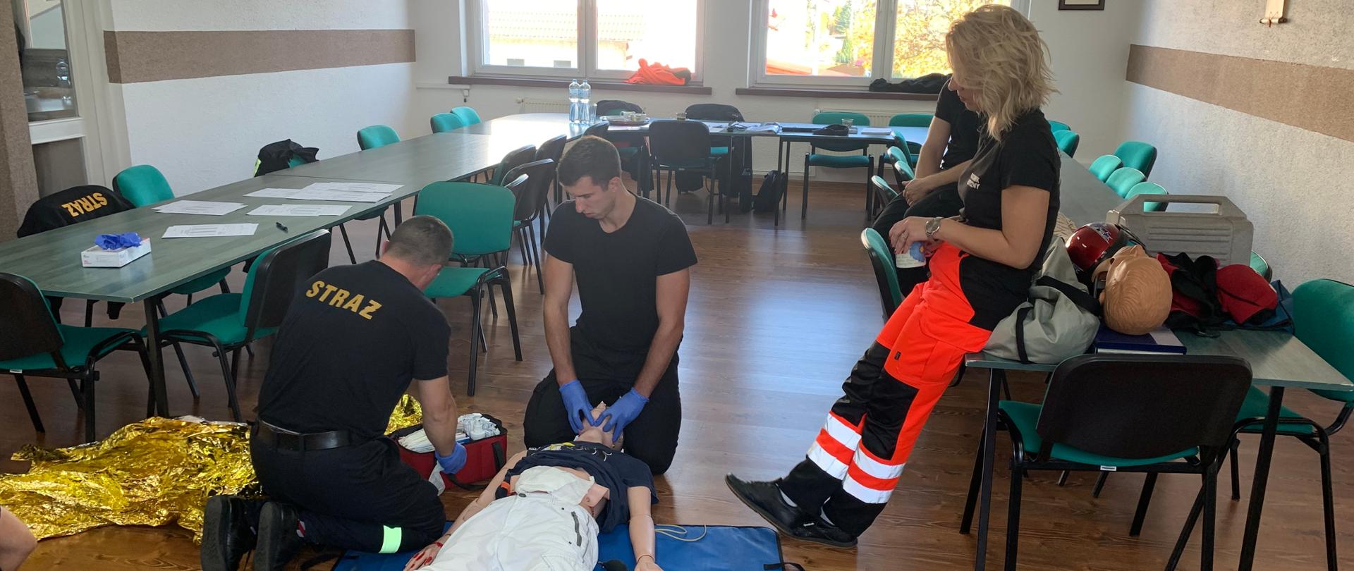 Na świetlicy Komendy stoły ustawione w podkowę. W środku podkowy na podłodze ułożono manekina z zamocowanymi sztucznymi ranami. manekin ubrany w jasne spodnie i koszulkę T-shirt. Dwóch strażaków udziela mu pomocy. Pierwszy klęczy za głową stabilizując ją kolanami, rękoma w rękawiczkach nitrylowych udrażnia drogi oddechowe. drugi ratownik zabezpiecza ranę brzucha. Po prawej oparta o stół członkini komisji egzaminacyjnej - w ubraniu ratownika medycznego w kolorze pomarańczowym. 