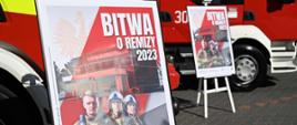 Dwa plakaty promocyjne akcji „Bitwa o Remizy 2023” na tle dwóch pojazdów pożarniczych