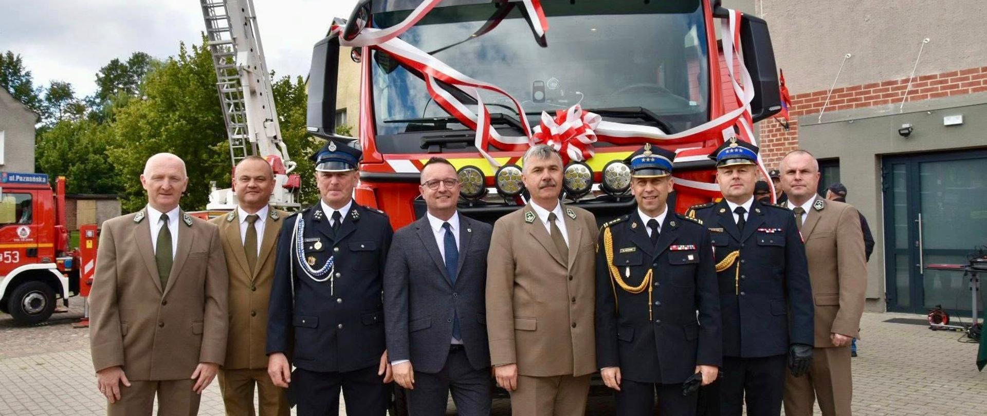 Na zdjęciu widzimy grupę ośmiu mężczyzn stojących przed czerwonym wozem strażackim udekorowanym biało-czerwonymi wstęgami i dużą kokardą. Mężczyźni ustawieni są w rzędzie, pozują do wspólnego zdjęcia. Część z nich ubrana jest w ciemno-granatowe mundury, a jeden w garnitur z krawatem. W tle widać drugi samochód strażacki oraz budynek z oknami.