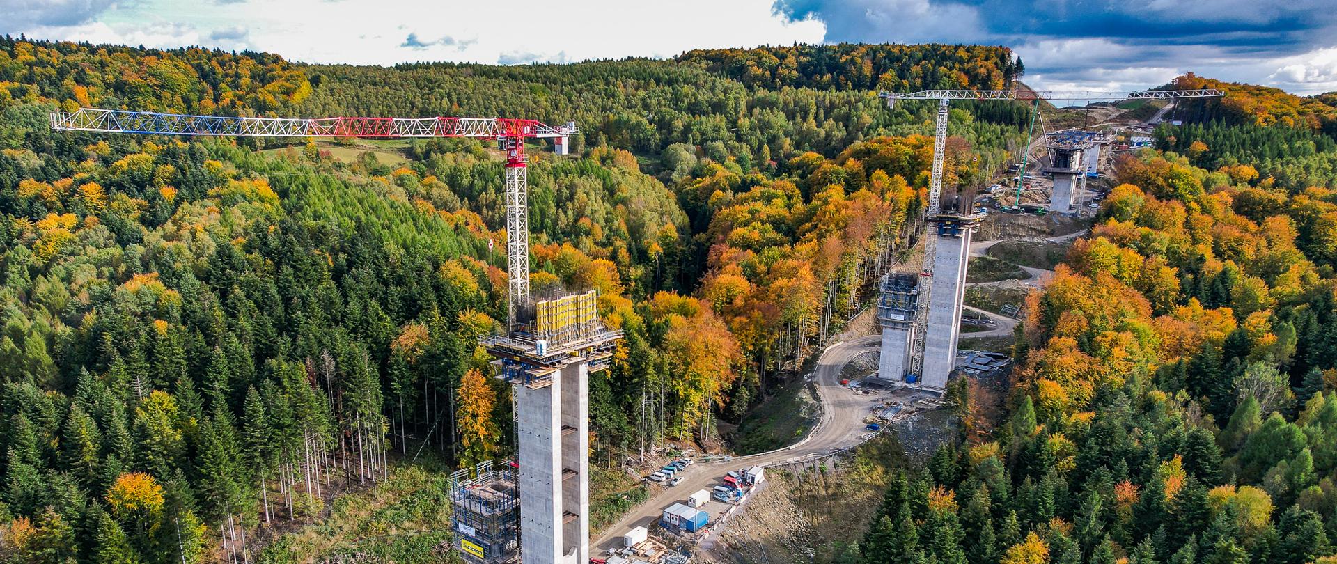 Zdjęcie przedstawia rozległy plac budowy estakady w górzystym, zalesionym terenie jesienią. Widać kilka bardzo wysokich, betonowych pylonów, na których mają powstać przęsła mostu. Na dwóch z nich stoją duże żurawie wieżowe. Wokół znajdują się drogi techniczne, maszyny i kontenery budowlane. Drzewa mają intensywne barwy jesieni — zielenie, żółcie i pomarańcze. Cała scena jest ujęta z góry, prawdopodobnie z drona.