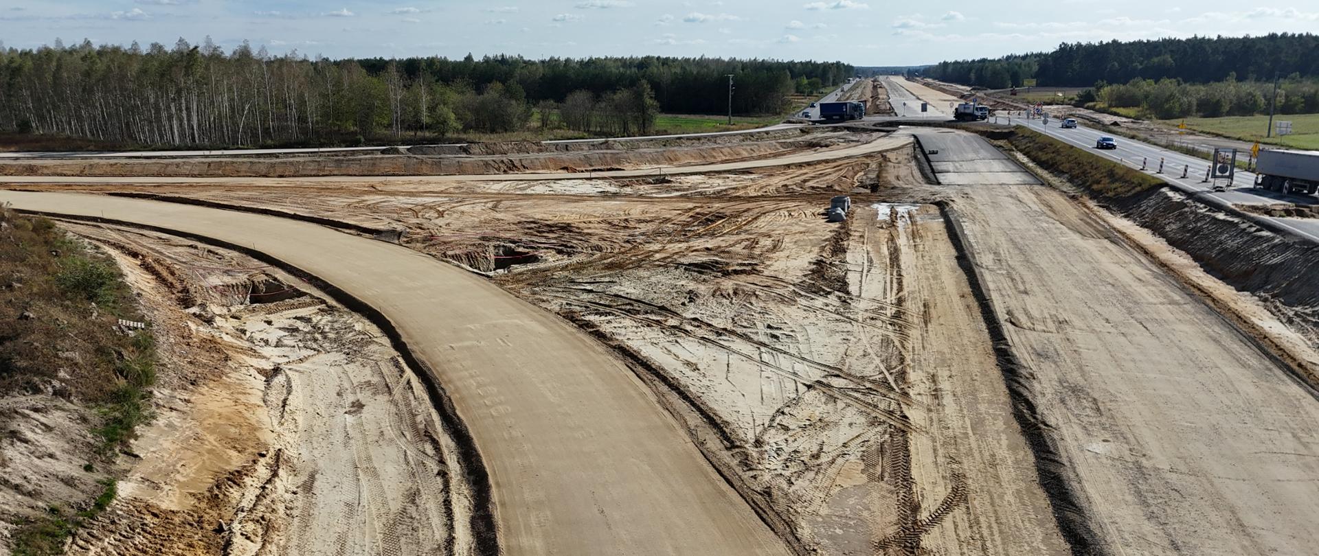 Zdjęcie przedstawia plac budowy drogi ekspresowej S19 pomiędzy Lublinem a Lubartowem. Na pierwszym planie widoczna jest szeroka, nieukończona jezdnia – pokryta warstwą ubitego kruszywa. W oddali widać objazd i samochody jadące drogą tymczasową. Teren jest otwarty, bez zabudowań, na horyzoncie widać las.