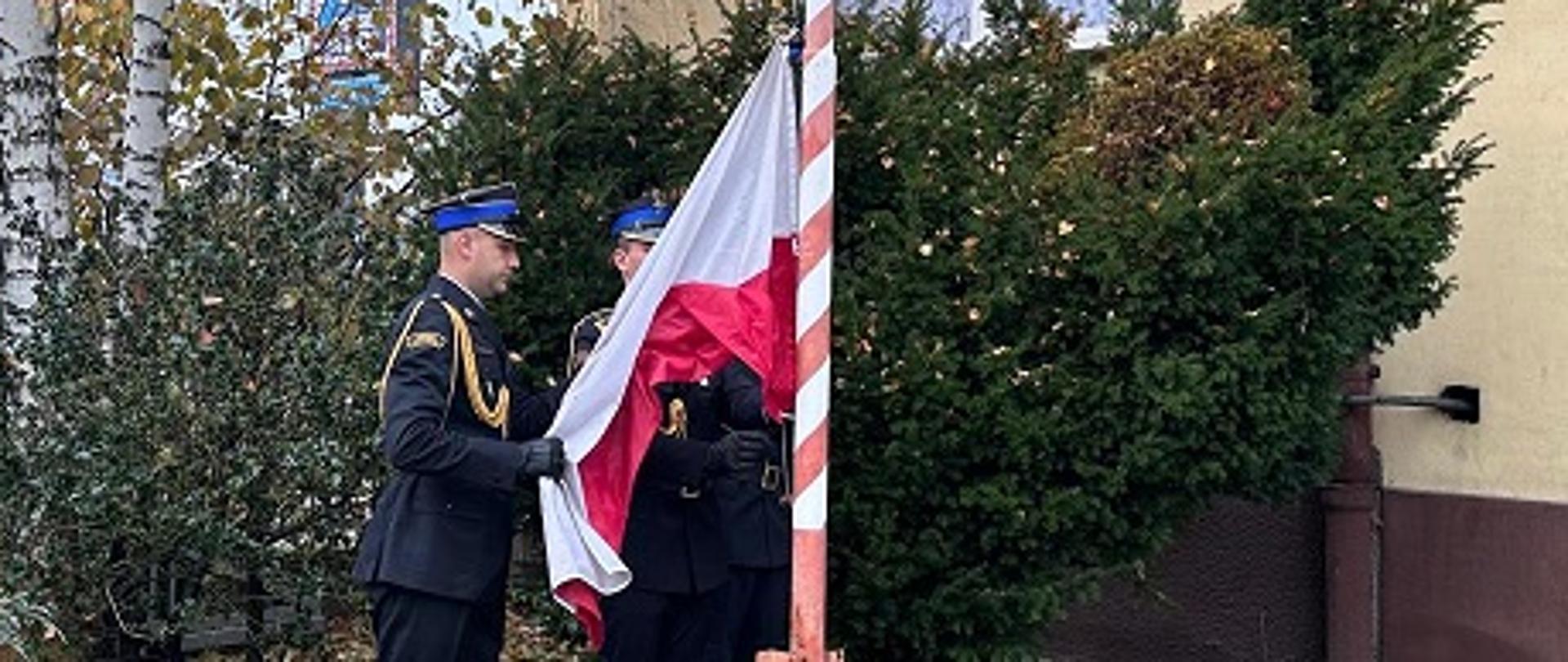 Na pierwszym planie maszt flagowy. Za nim stoi poczet flagowy, który przymocowuje flagę państwową do linki nośnej, w celu wciągnięcia flagi na maszt. 