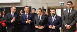 Minister Czarnek i czterech mężczyzn w garniturach stoją w rzędzie i biją brawo, za nimi grupa osób w ceremonialnych czarno-czerwonych strojach.