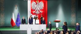 Uroczysta ceremonia z udziałem najwyższych władz państwowych. Przed podium stoją przedstawiciele rządu i wojska, a w tle zawieszone są flagi Polski i Unii Europejskiej oraz duży orzeł biały na czerwonym tle. Po lewej stronie widać krzyż, a po obu stronach – bogato zdobione zielone tło