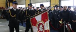 Trzech strażaków maszeruje ze sztandarem podczas uroczystej zbiórki, w tle zaproszeni goście