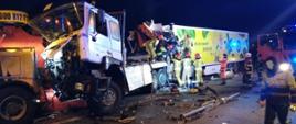 Pora nocna. Na zdjęciu widać wypadek z udziałem samochodów ciężarowych. Samochody są ze sobą szczepione. Przy kabinie trzeciego w kolejności pojazdu znajduje się grupa strażaków udzielając pomocy osobie poszkodowanej. Z prawej strony stoi samochód ratowniczo-gaśniczy