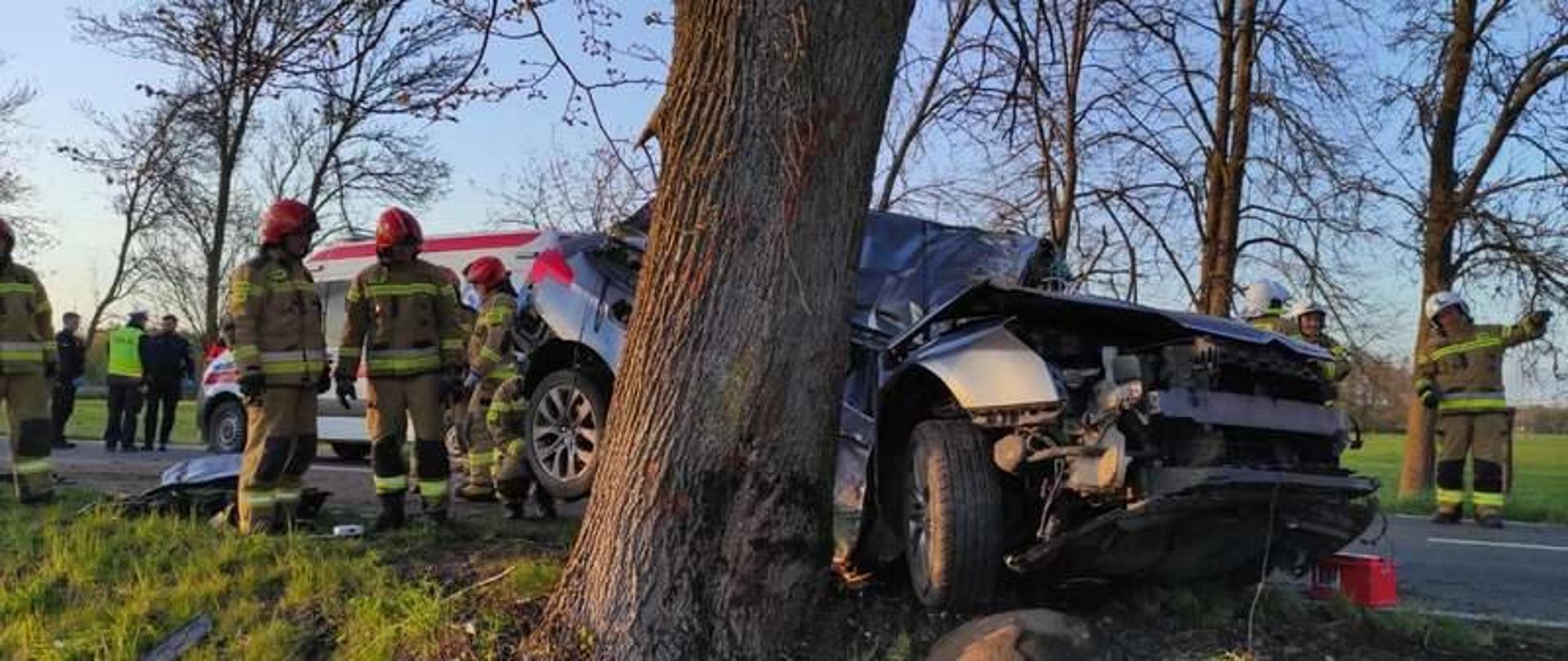 Na zdjęciu rozbite, mocno uszkodzone auto osobowe które uderzyło bokiem w drzewo. Na drodze strażacy.