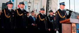 Widok z przodu. Trybuna honorowa, na której stoją goście zaproszeni na uroczystość. Na pierwszym planie przemawia zastępca małopolskiego komendanta wojewódzkiego PSP.