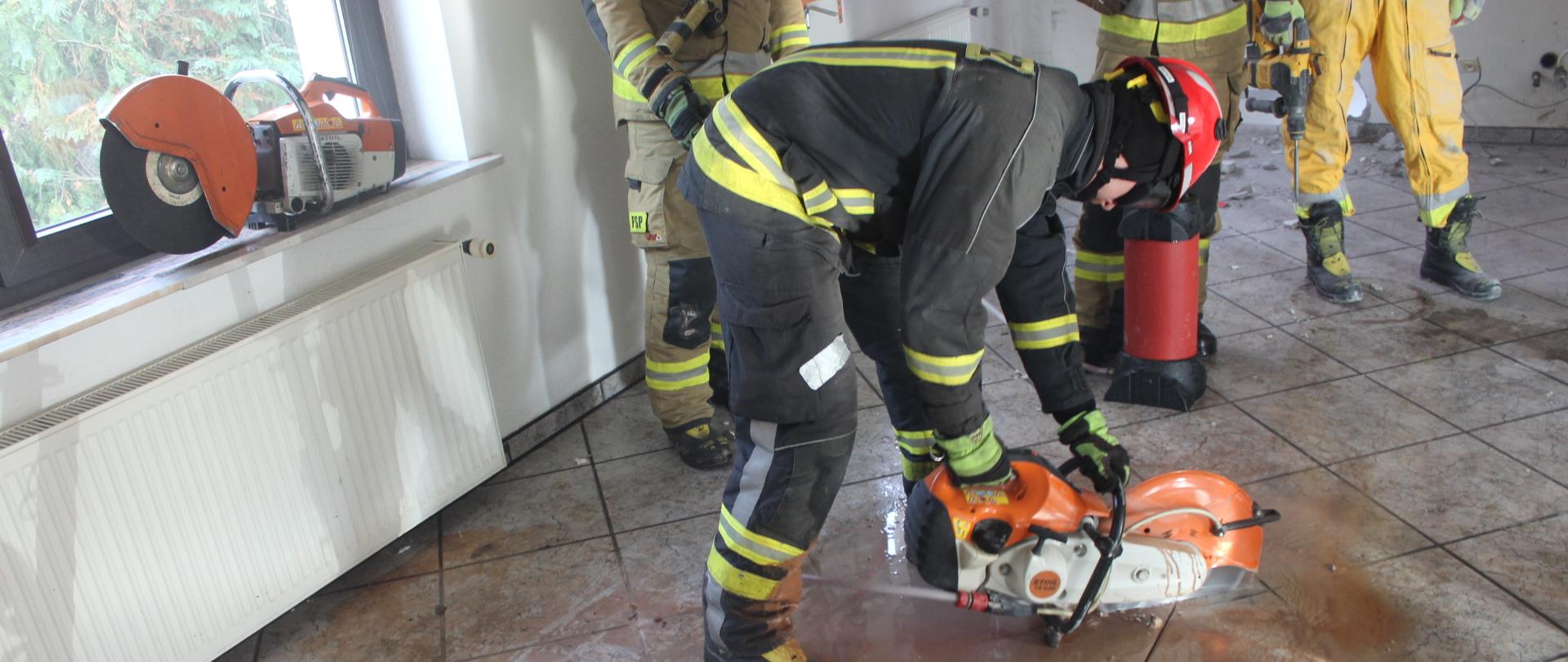 Strażak przy pomocy przecinarki do stali i betonu wykonuje nacięcie podłogi w budynku jednorodzinnym. 