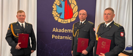 Trzech funkcjonariuszy Państwowej Straży Pożarnej z dyplomami pozuje do zdjęcia na tle ścianki Akademii Pożarniczej.