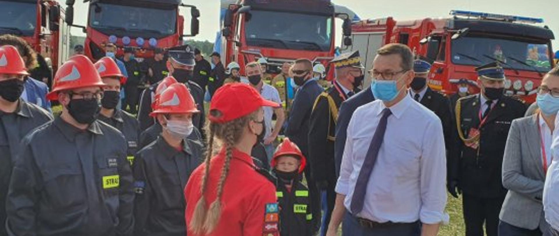 Premier Polski rozmawia z młodzieżową drużyną pożarniczą. W tle strażacy i pojazdy pożarnicze.