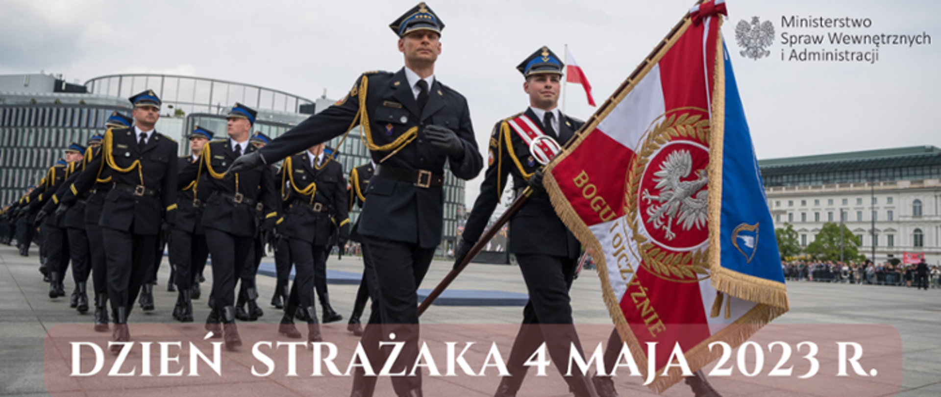 Zdjęcie wykonane na w Warszawie na Placu Marszałka Józefa Piłsudskiego. Na zdjęciu widać uroczystą defiladę pododdziałów Państwowej Straży Pożarnej z okazji obchodów Dnia Strażaka. Na pierwszym planie widać poczet sztandarowy. Poczet tworzy trzech funkcjonariuszy ubranych w mundury galowe. Środkowy z nich w rękach trzyma sztandar. Dodatkowo jego mundur przepasany jest biało – czerwoną wstęgą. Za pocztem sztandarowym maszerują w czwórkach pozostali funkcjonariusze również ubrani w mundury galowe. W oddali widać zabudowania okalające plac, na którym odbywa się uroczystość. 