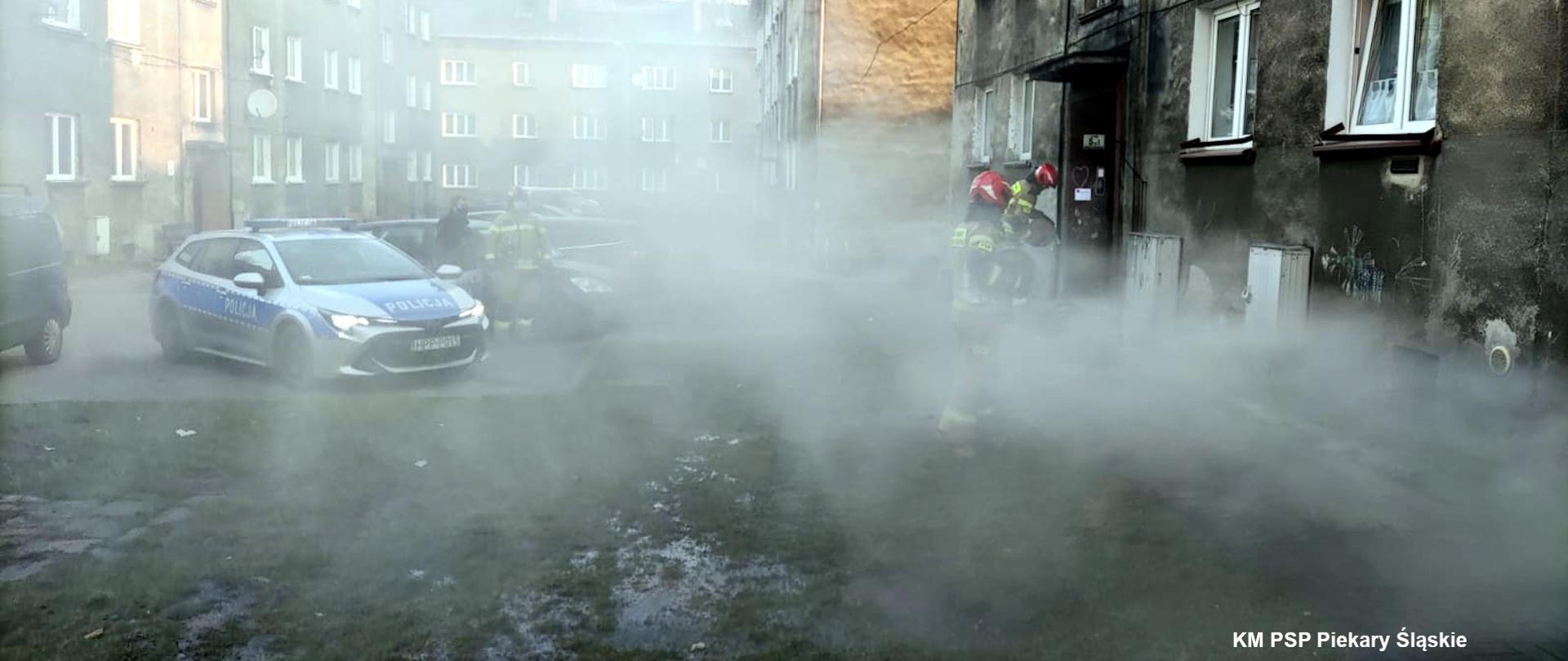 Zdjęcie przedstawia zadymiona przestrzeń przed budynkiem w którym doszło do pożaru. Na zdjęciu widoczni są również strażacy oraz radiowóz policji.
