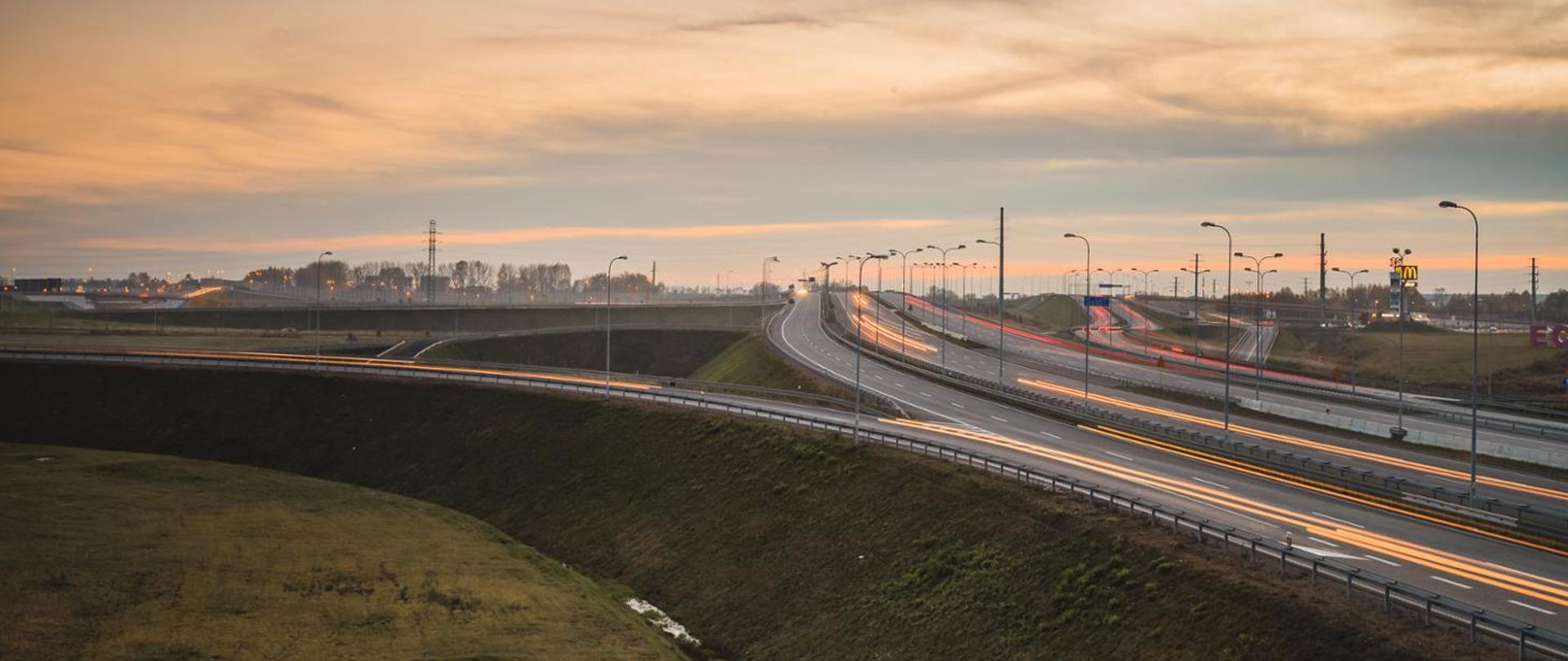 Fotografia węzła autostradowego o zachodzie słońca. Widoczne wydłużone ślady świateł samochodowych. Niebo zachmurzone.