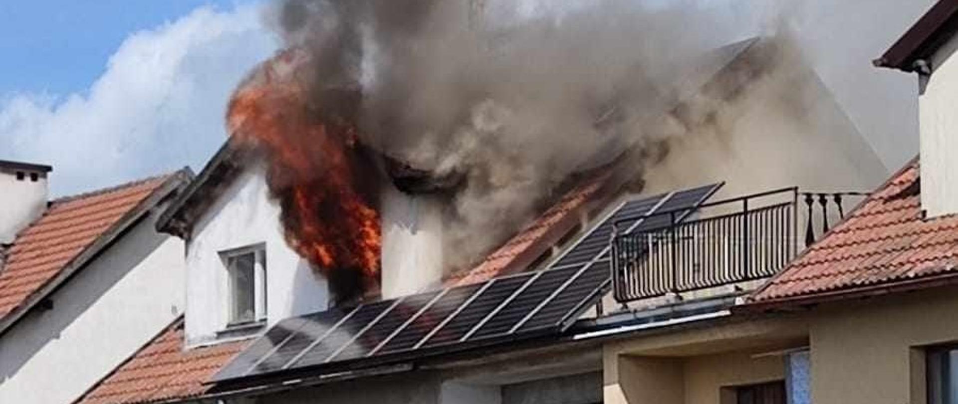 Zdjęcie przedstawia języki ognia i kłęby dymu wydobywające się z okna na poddaszu budynku mieszkalnego oraz samochód gaśniczy stojący przed budynkiem.