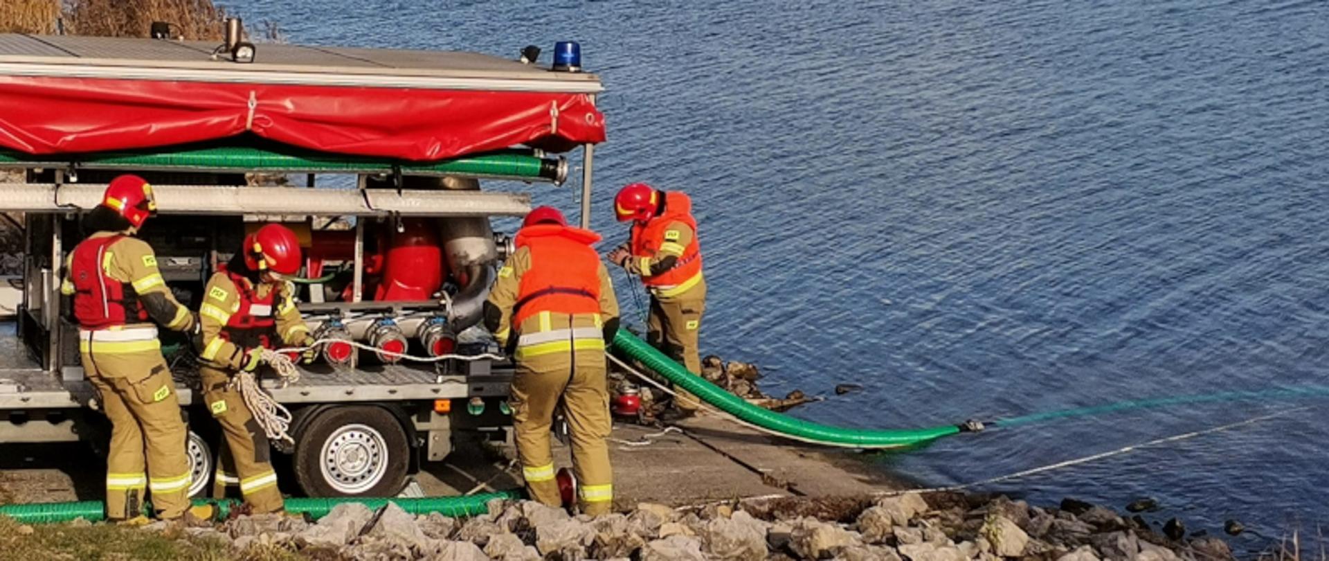 Na zdjęciu czterech strażaków ubranych w ubrania specjalne oraz czerwone kamizelki ratunkowe odpompowujący wodę do akwenu wodnego z motopompy 