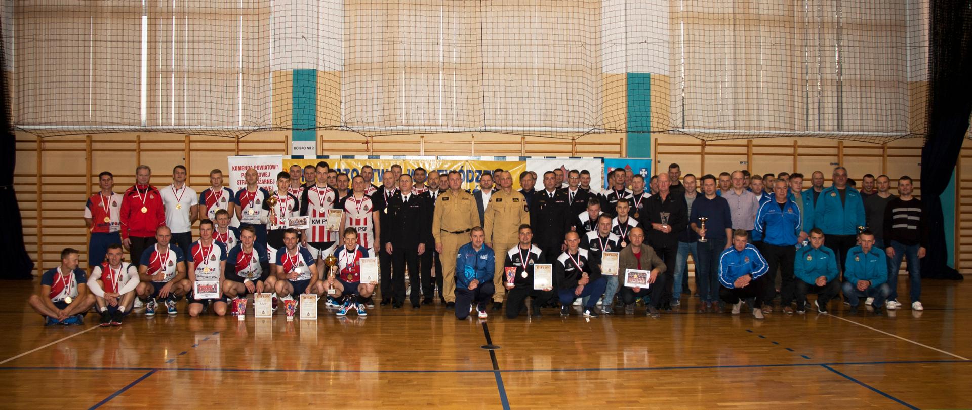Zdjęcie grupowe wszystkich uczestników zawodów – łącznie kilkadziesiąt osób. Członkowie drużyn w strojach sportowych i w ubraniach cywilnych – część stoi, część kuca. Część osób z medalami, dyplomami i pucharami. Pomiędzy zawodnikami, w środki kilkunastu strażaków, m.in. podkarpacki komendant wojewódzki PSP oraz komendanci powiatowi i miejscy PSP w umundurowaniu. Zdjęcie na sali gimnastycznej.