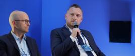 Wiceminister funduszy i polityki regionalnej Konrad Wojnarowski siedzi w fotelu i przemawia do mikrofonu, po jego prawej stronie siedzi inny uczestnik dyskusji