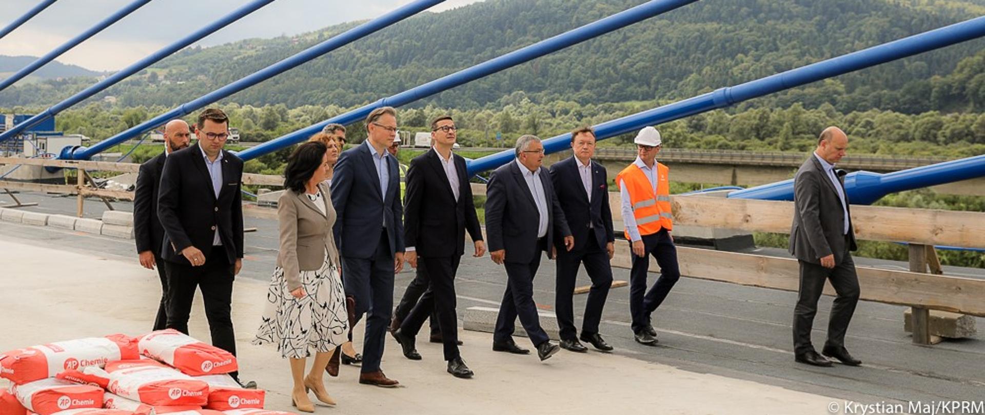 Premier z ministrem infrastruktury i samorządowcami zwiedzają teren budowy mostu.