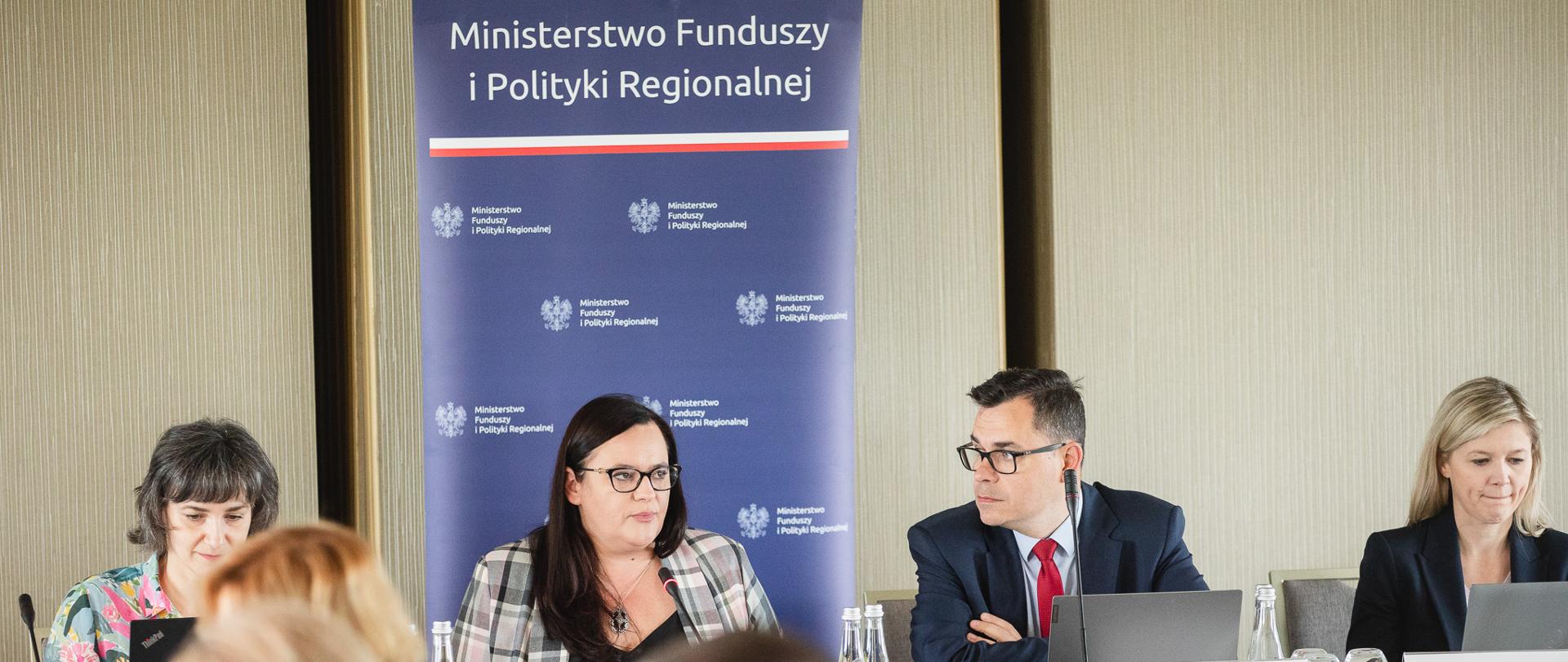 Na pierwszym planie 3 osoby. W tle siedzą 4 osoby za stołem konferencyjnym.