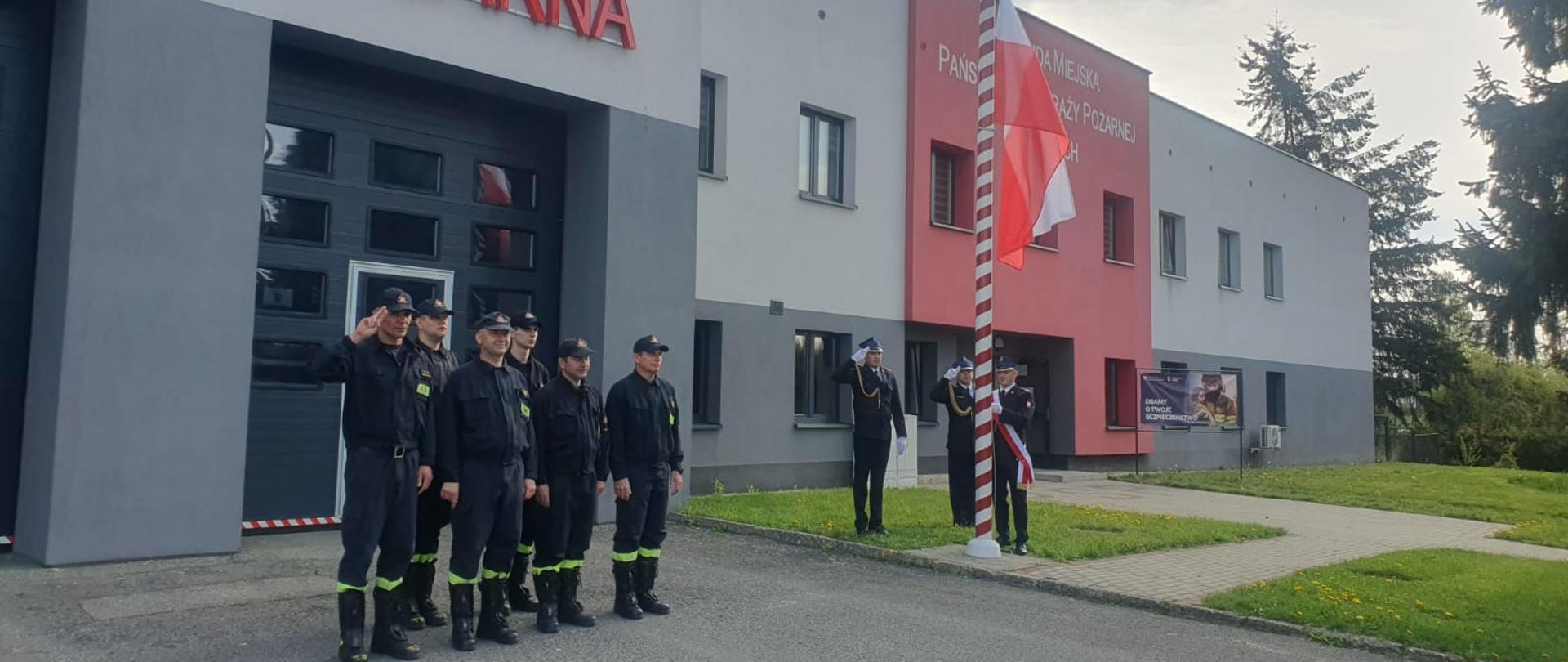 Strażacy stojący w dwuszeregu na tle budynku Komendy jeden z nich salutuje, trzech strażaków stoi przy maszcie flagowym, jeden mocuje flagę, dwaj salutują.