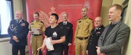 Komendant Miejski w mundurze wyjściowym. Obok niego strażacy w mundurach i pan w marynarce Chłopak w czarnej koszulce trzyma dyplom i torbę sportową w rękach przed dorosłymi. Za nimi czerwona ścianka firmowa z dużym orłem białym, napisem Państwowa Straż Pożarna i logo PSP.