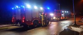 Widok na ulicę. Zdjęcie wykonane w nocy. Wzdłuż drogi stoją auta strażackie. W tle widoczne domki jednorodzinne.