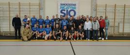 Zdjęcie przedstawia strażaków w strojach sportowych oraz Komendanta Powiatowego w mundurze piaskowym na parkiecie hali sportowej
