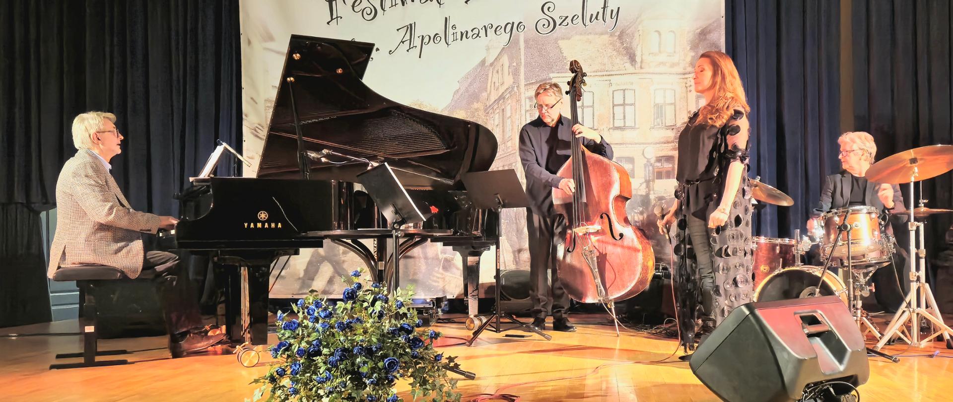 zespół złożony z czterech muzyków podczas występu na scenie, przed sceną bukiet kwiatów, w tle baner konkursowy i granatowa kotara