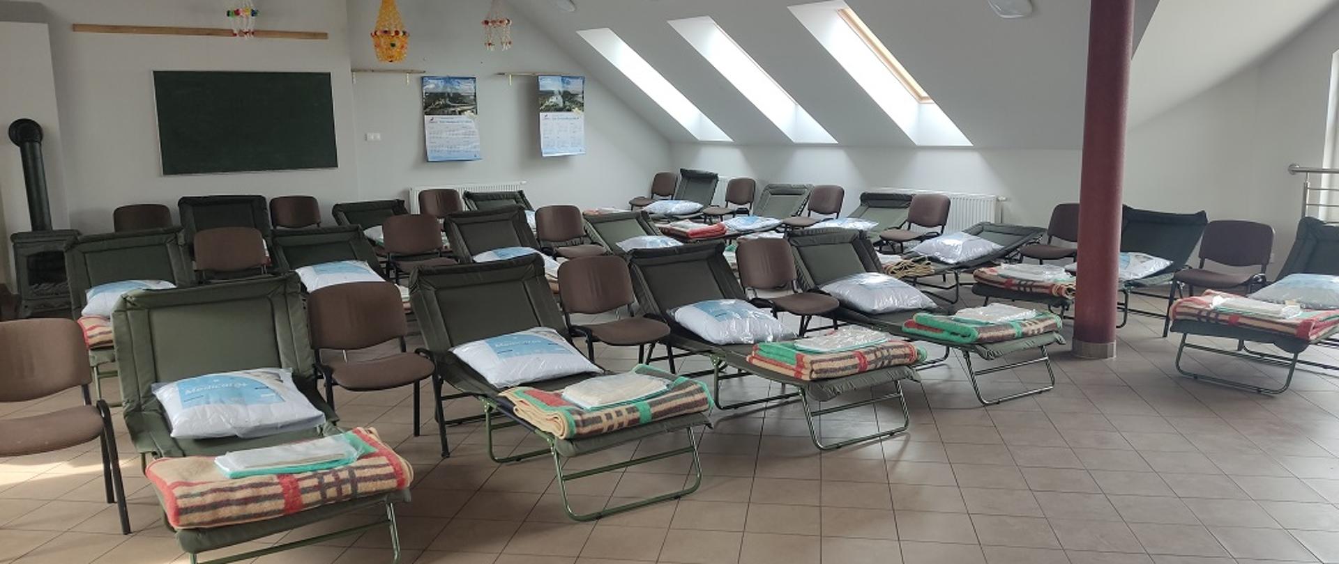 Na zdjęciu widoczne są rozstawione łóżka polowe na sali. Na łóżkach znajduje się pościel oraz poduszki. Ściany pomieszczenia są pomalowane w kolorze białym. Na jednej ze ścian widać zawieszony telewizor oraz ścienne kalendarze. Przy ścianie widoczny jest również kominek