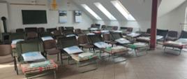 Na zdjęciu widoczne są rozstawione łóżka polowe na sali. Na łóżkach znajduje się pościel oraz poduszki. Ściany pomieszczenia są pomalowane w kolorze białym. Na jednej ze ścian widać zawieszony telewizor oraz ścienne kalendarze. Przy ścianie widoczny jest również kominek