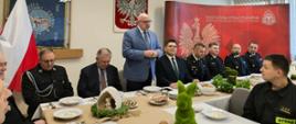 Przy stołach na których znajdują się dekoracje świąteczne i talerze, siedzą strażacy i zaproszenie goście. Strażacy ubrani są w mundury galowe zaproszeni goście odświętnie w garniturach. Jeden z zaproszonych gości w postawie stojącej przemawia. W tle logo Państwowej Straży Pożarnej oraz godło Polski i flagi