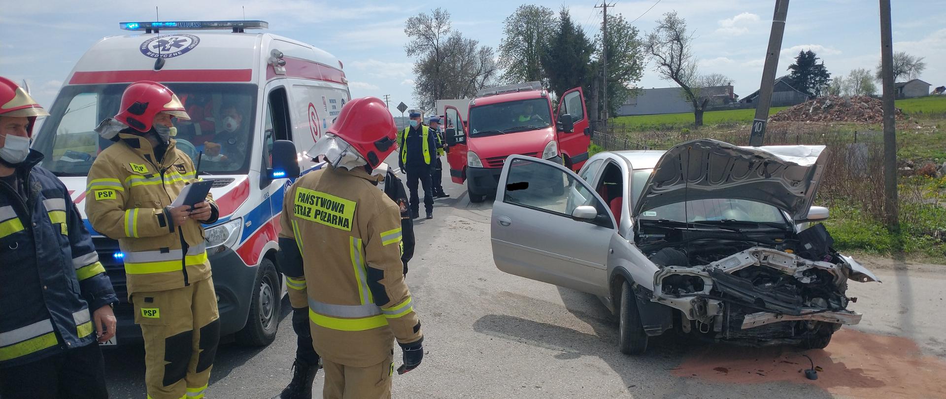 Zdjęcie przedstawia strażaków stojących obok karetki Zespołu Ratownictwa
Medycznego oraz uszkodzony samochód.
