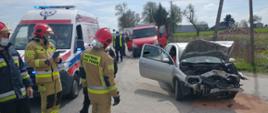 Zdjęcie przedstawia strażaków stojących obok karetki Zespołu Ratownictwa
Medycznego oraz uszkodzony samochód.
