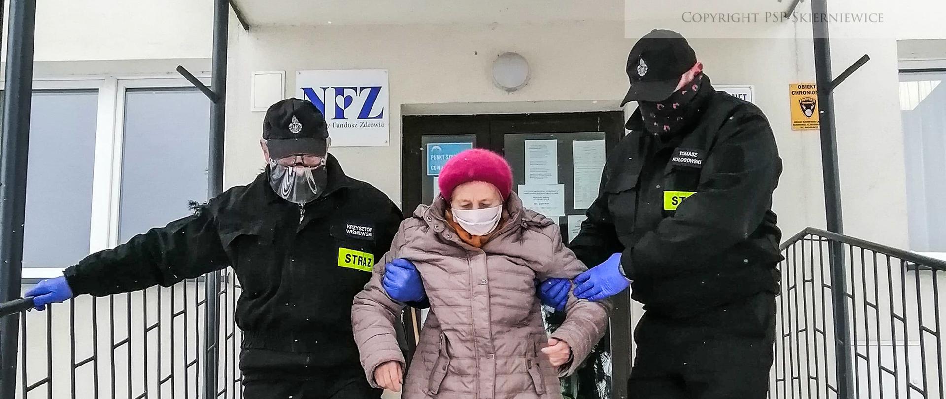 Druhowie OSP pomagają starszej osobie w zejściu ze schodów.