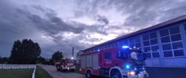 Na zdjęciu pojazdy ratowniczo-gaśnicze. Pojazdy mają włączone światła ostrzegawcze. Wieczór. Duże zachmurzenie. Na drugim planie budynek sali gimnastycznej. Auta stoją na drodze.