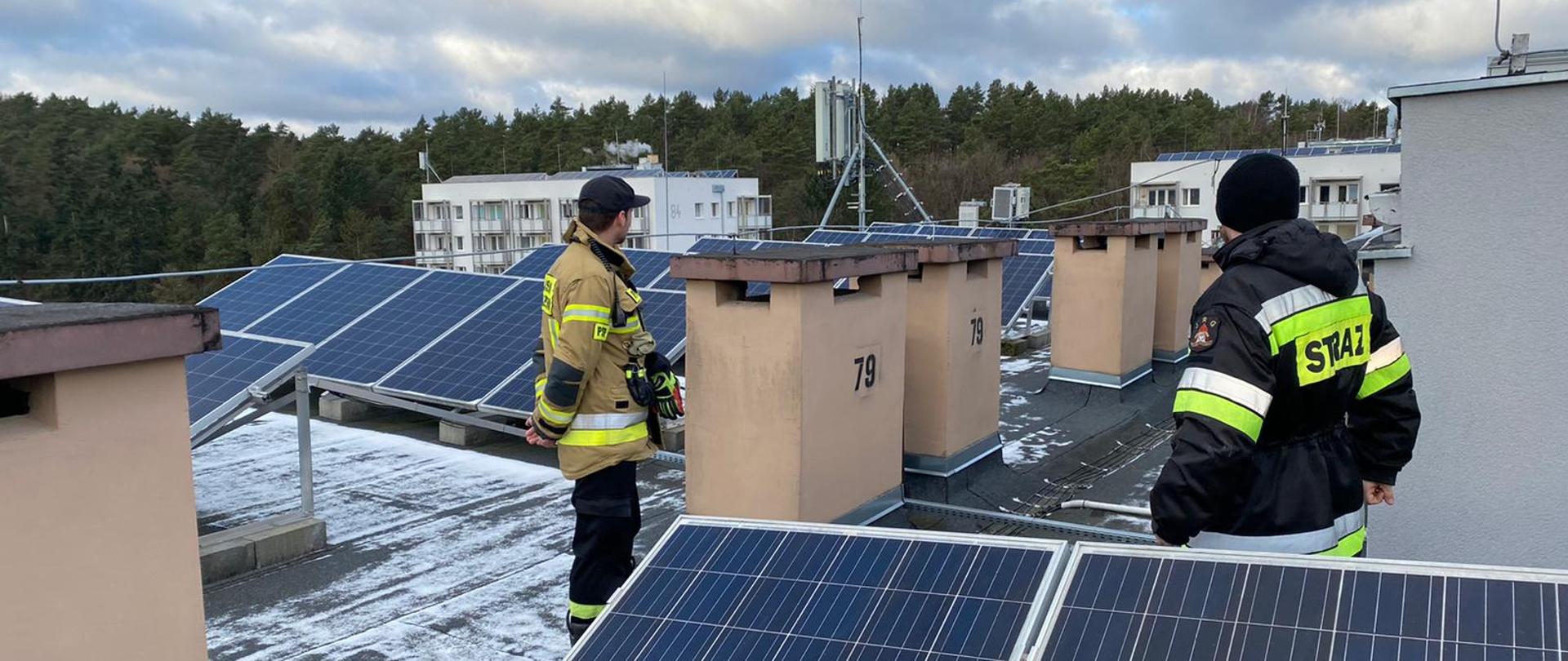 Dwóch strażaków sprawdza na dachu budynku wysokiego instalacje fotowiltaiczne zamontowane w kilku szeregach