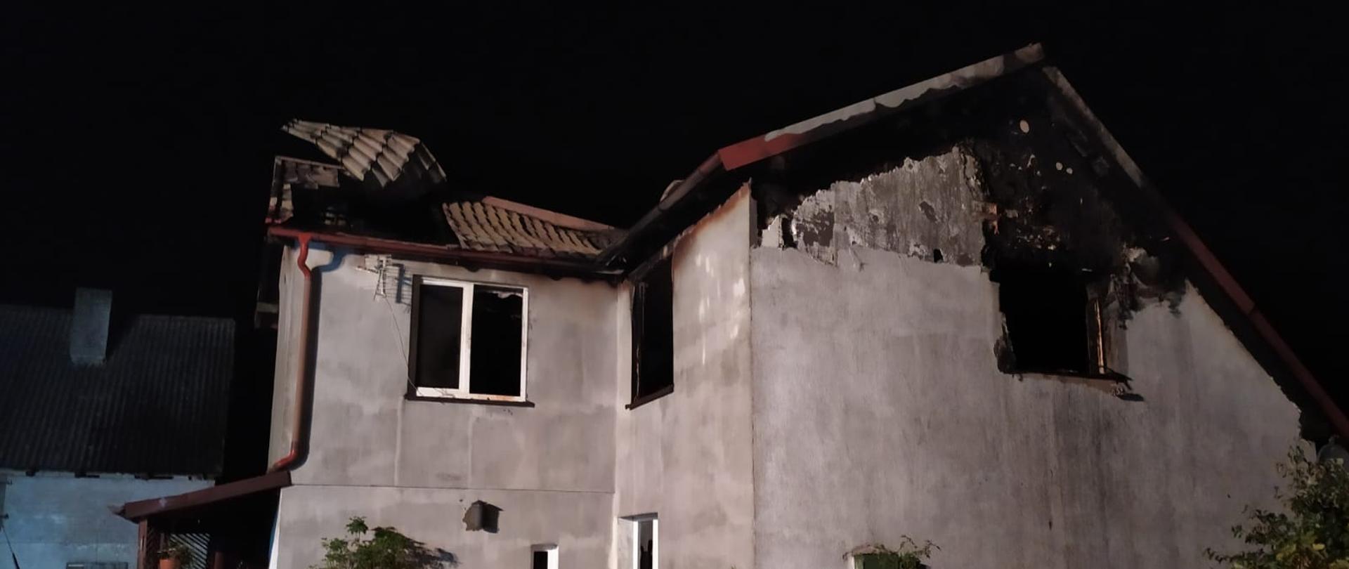 Zdjęcie przedstawia budynek domku jednorodzinnego, wykonano to zdjęcie nocą. Za budynkiem widać budynek gospodarczy, po lewej stronie widnieje kawałek samochodu osobowego koloru czerwonego, po prawej zaś kawałek ogrodzenia oraz ogródka na dachu widać pofalowaną blachę, która pod naciskiem ciepła od pożaru się pofalowała cała góra budynku jest częściowo wypalona 