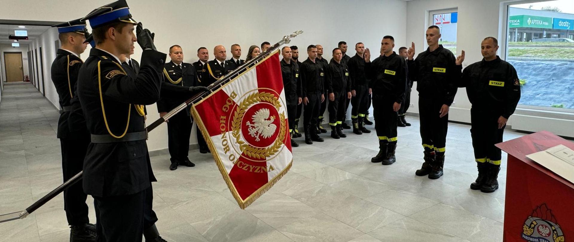 Na zdjęciu znajduje się poczet sztandarowy KM PSP w Elblągu wraz ze sztandarem komendy, trzech strażaków składających ślubowanie oraz pododdział funkcjonariuszy KM PSP w Elblągu. Nowo przyjęci strażacy składają ślubowanie w sali szkoleniowej komendy. 