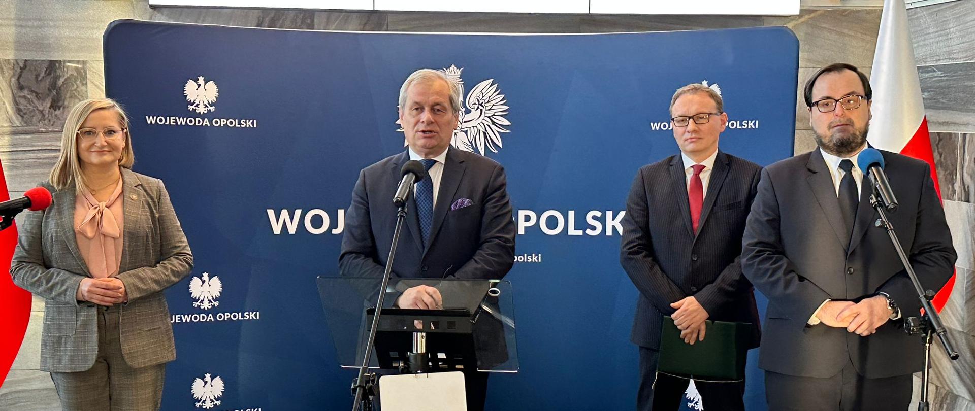 4 osoby stoją przy mikrofonach, 3 mężczyzn w garniturach i jedna kobieta w garniturze. Za nimi znajduje się granatowa ścianka z napisami województwo opolskie