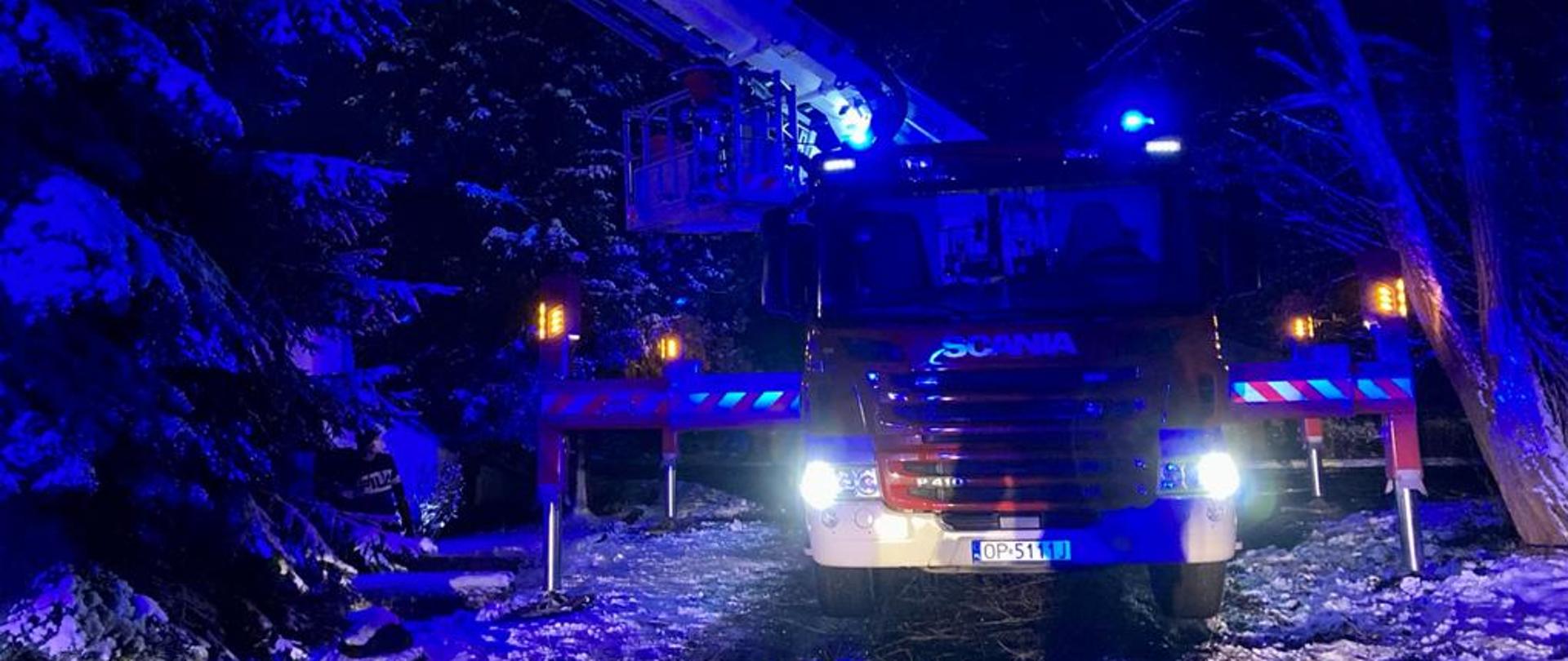 Zdjęcie przedstawia ustawiony podnośnik straży pożarnej w porze nocnej rozstawiający się pomiędzy drzewami i krzewami. Widok podnośnika marki Scania koloru czerwonego od przodu pojazdu.
