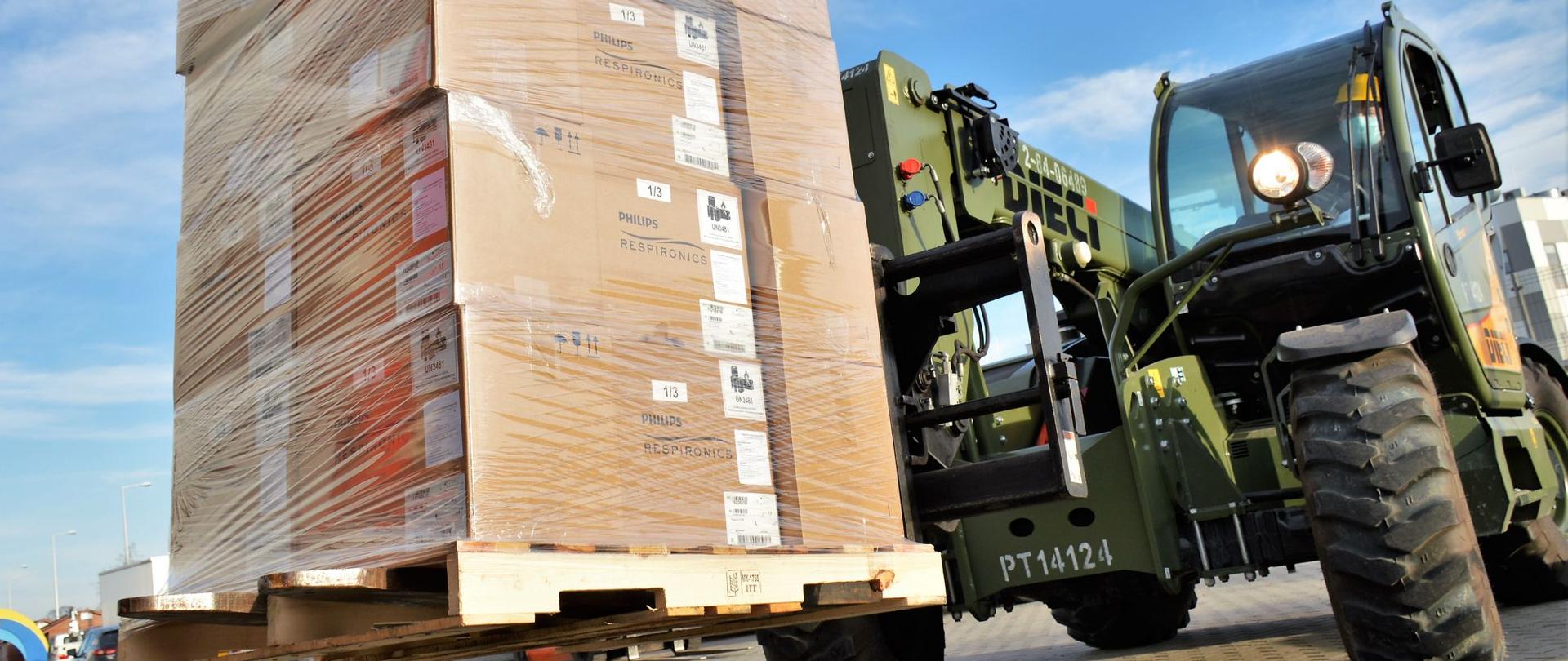 Na zdjęciu widać pojazd dostawczy, przypominający traktor, wożący paletę z paczkami ze środkami ochrony osobistej. Pojazd jest w kolorze zielonym. W tle widać budynek szpitala tymczasowego w Opolu. 