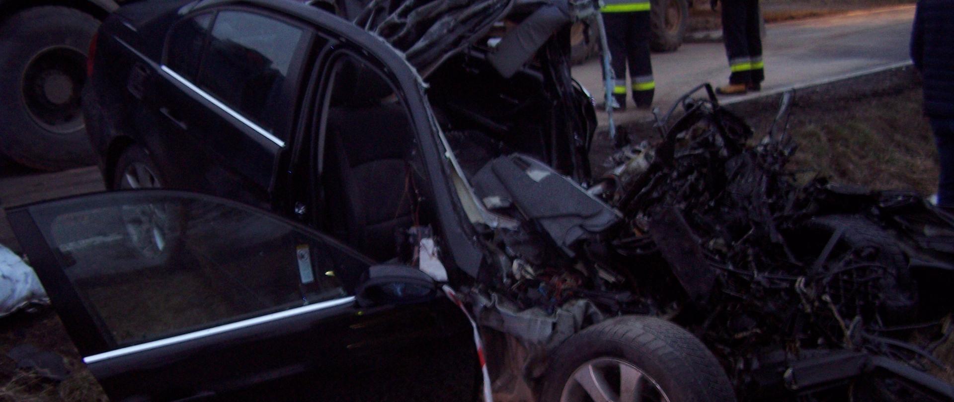 Samochód osobowy znajduje się na poboczu jezdni. Zmiażdżony cały przód pojazdu oraz przedział pasażerski aż do tylnych siedzeń. 