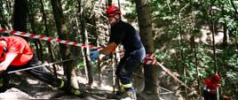 Strażacy w lesie przy użyciu liny wyciągają poszkodowanego. W tle widoczna taśma ostrzegawcza.